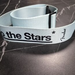Světle modrý textilní popruh k hudebnímu nástroji s černými plastovými prvky. Na popruhu je černý nápis „to the Stars“ a logo „Fender“. Povrch popruh…
