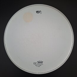 Bílá kruhová bubenová koží s bílým plastovým okrajem. V horní části je černý nápis 15" a v dolní části logo s korunkou a texty REMO, UT DRUM HEAD, MA…