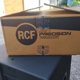Kartonová krabice s nápisy RCF PRECISION TRANSDUCERS položená na černém reproduktorovém boxu. Krabice je z hnědého kartonu s černým pruhem a je slepe…