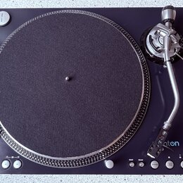 Černý gramofon s kulatým talířem potaženým tmavě šedou plstí a stříbrným vlnitým okrajem. Na raménku je viditelné logo "aton" s modrým symbolem. Ovlá…