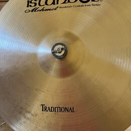 Zlatý kovový činel s viditelným opracováním povrchu. V horní části je černý nápis istanbul® a pod ním kurzívou Mehmet Handmade Cymbals from Turkey. V…