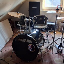 Černá bicí souprava značky YAMAHA s chromovaným hardwarem. Na basovém bubně je bílý nápis YAMAHA s logem a v dolní části malý kulatý štít. V pozadí s…