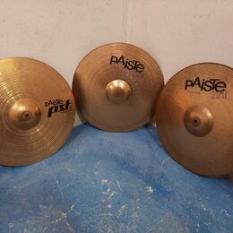 Tři mosazné činely opřené o bílou zeď na modré podlaze. Nejlevý činel vlevo nese nápisy paiste PST II a 18" CRASH RIDE. Prostřední činel má nápisy pA…