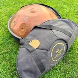 Na fotografii je kovový nástroj typu handpan v měděné barvě, umístěný v černé přepravce. Na povrchu nástroje je nápis „Handpan Design Sela“. Před nás…