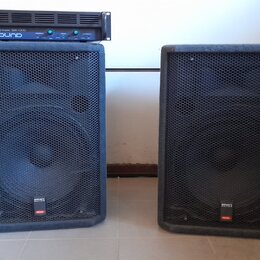 Dva černé reproduktorové boxy s kovovými ochrannými mřížkami. Na spodní části obou boxů je malý štítek s nápisy „SOUND SYSTEM“ a „PROEL“. Na levém re…