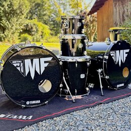 Černá bicí souprava složená z dvou basových bubnů, jednoho tom-tomu a jednoho floor tomu. Na předních blanách basových bubnů je bílý nápis METAL FACT…