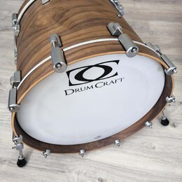 Kytlicový basový buben s dřevěným korpusem v hnědé barvě a bílým exponovaným koženem. Na koženě je černý nápis DRUM CRAFT a nad ním grafické logo. Bu…