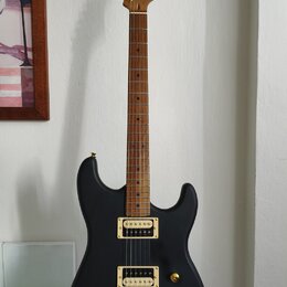 Černá elektrická kytara s matným povrchem a zlatým hardwarem. Na hlavě hmatníku je viditelné logo "Cort". Nástroj má dva humbuckery a zlatý most. Kyt…