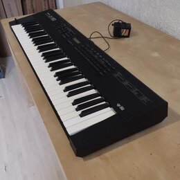 Černý digitální klavír s klávesnicí umístěný na světlém dřevěném stole. Na horním panelu je vlevo nápis "Roland" a vedle něj modelové označení "JV-10…