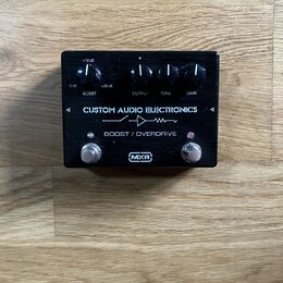 Černý efekt pedál s nápisy CUSTOM AUDIO ELECTRONICS a BOOST / OVERDRIVE. Na horní části jsou čtyři ovládací knoflíky s popisky BOOST (s rozsahem 0 dB…