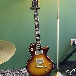 Elektrická kytara s trupem v barvě sunburst a tmavým hmatníkem s trapezoidními vložkami. Na hlavě je bílý nápis Gibson a kurzivní nápis Les Paul. Nás…