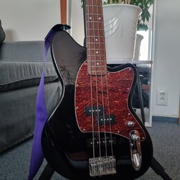 Černá elektrická basová kytara s tmavě hnědým hmatníkem. Nástroj má červený pickguard s texturou imitující dřevo a dva snímače. Na těle není vidné žá…