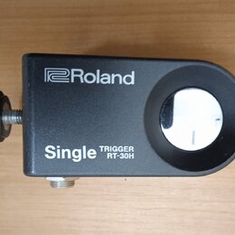 Černý trigger s nápisy Roland a Single TRIGGER RT-30H. Zařízení má obdélníkový tvar s vykrouženým otvorem a je vybaveno šroubovacím knoflíkem. Povrch…