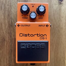 Oranžový efekt pedál s černým spínacím tlačítkem. Na zařízení jsou nápisy Distortion DS-1, BOSS, TONE, LEVEL, DIST, OUTPUT, INPUT a CHECK. Pedál má t…