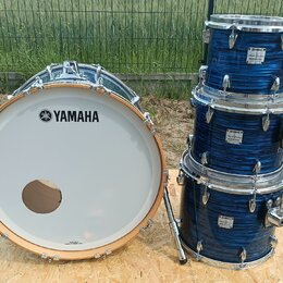 Modrá bicí souprava s kovovým hardwarem. Na bílém bendášu basového bubnu je černé logo a nápis YAMAHA. Na třech tom-tamech jsou viditelné bílé štítky…