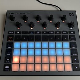 Šedý sampler s nápisem CIRCUIT RHYTHM. Zařízení obsahuje několik černých otočných ovladačů a mřížku podsvícených padů v modré a oranžové barvě. Na po…