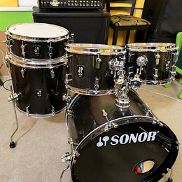 Černá bicí souprava značky SONOR. Na basovém bubně je bílé logo SONOR, na ostatních bubnech jsou viditelné štítky s nápisem ACK. V pozadí je kytarový…