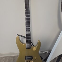 Zlatá elektrická kytara s dvojcutem, umístěná na černém stojanu. Na hlavě hmatníku je čitelné logo LTD. Nástroj má dva humbuckery, zlatý hardware a T…