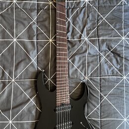 Černá elektrická kytara s tmavým hmatníkem a dvěma humbuckery. Na hlavě je bílý nápis Ibanez. Nástroj vykazuje známky běžného používání, jsou vidět d…