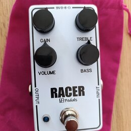 Bílý efekt pedál s nápisem RACER a logem M pedals. Na horní straně jsou čtyři černé oválné ovládací knoflíky s popisky GAIN, TREBLE, VOLUME a BASS. V…