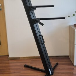 Černý kovový stojan na klávesové nástroje s dvěma vodorovnými rameny. Stojan má šikmý hlavní sloup s integrovanou rukojetí pro přenos a křížovou zákl…