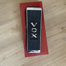 Černý kytarový wah-pedál s chromovanými okraji, umístěný na červené krabici. Uprostřed je nápis VOX a po stranách jsou texty „2 X OHMS 10W“ a „R.S.A.…