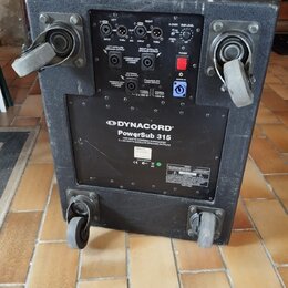 Černý aktivní subwoofer v texturovaném krytu s dvěma madly a dvěma kolečky. Na zadním panelu je ovládací sekce s konektory, potenciometry a červeným…