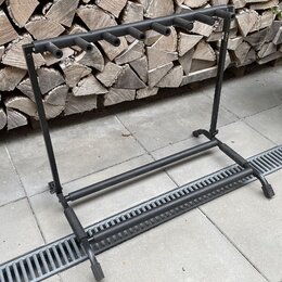 Černý kovový stojan na několik kytar s vodorovnou horní lištou vybavenou pěti držáky na krky. Konstrukce tvoří rám s dvěma svislými nohami a spodní o…