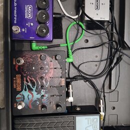 Sestava efektů v černém kufru. Obsahuje fialový pedál s nápisy MXR CUSTOM SHOP sub machine, stříbrný napájecí adaptér s logem MXR a černý pedál s náp…