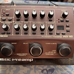 Hnědý kytarový předzesilovač s nápisy BOSS a Acoustic Preamp AD-10. Zařízení má v horní části řadu ovládacích knoflíků, přepínačů a malý digitální di…
