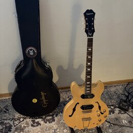 Světle hnědá polodubová elektrická kytara s dvěma humbuckery a f-otvory, umístěná na stojanu. Na hlavě hmatníku je bílý nápis Epiphone a logo v podob…