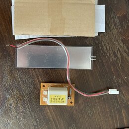 Fotografie zobrazuje elektronickou součástku s hliníkovým chladičem a přiloženou hnědou papírovou obálkou. Na oranžovém plošném spoji je bílý čtverco…
