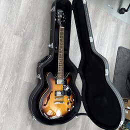 Polodupatá elektrická kytara s barevným přechodem od žluté po tmavě hnědou v černém tvrdém pouzdře. Na hlavě nástroje je bílý nápis Epiphone. Kytara…