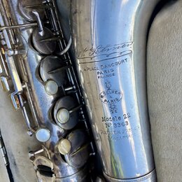 Detail stříbrného saxofonu s viditelným opotřebením povrchu a drobnými škrábanci. Na trubici jsou vyryty nápisy „H. Selmer“, „4. PLACE DANCOURT PARIS…