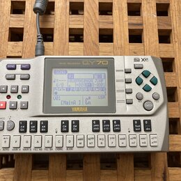 Stříbrný sekvencer Yamaha QY70 s LCD displejem. Na horní části je nápis MUSIC SEQUENCER a logo QY70, v pravém horním rohu jsou značky general MIDI a…