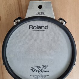 Kruhový elektronický trigger pad v šedé a černé barvě s kovovým uchytem. Na horní části je nápis PD-85 DUAL TRIGGER PAD a na membráně logo Roland s d…