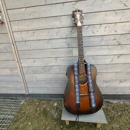 Akustická kytara s tmavě hnědým, přechodovým povrchem (sunburst) a modrým vzorovaným popruhem. Na hlavě nástroje je bílé logo Epiphone. Kytara stojí…