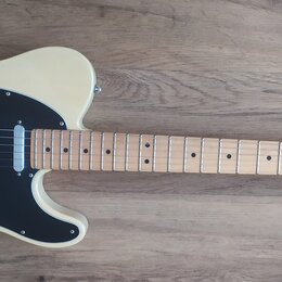 Elektrická kytara s krémovým korpusem, černým pickguardem a světle hnědým hmatníkem. Na hlavě krku je nápis Fender TELECASTER. Nástroj vykazuje čistý…