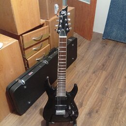 Černá elektrická kytara s špičatou hlavou a dvěma humbuckery, umístěná na stojanu. Na hlavě je bílé logo Ltd. Kytara má tmavý hmatník s bílými poloho…