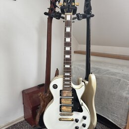Na fotografii jsou tři elektrické kytary v držáku. V popředí je bílá kytara typu Les Paul s černým pickguardem a dvěma humbuckery, na hlavě má nápis…