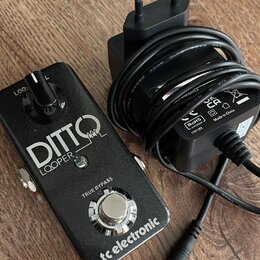 Černý kytarový pedál s nápisy DITTO LOOPER, TRUE BYPASS a tc electronic. Vedle pedálu je černý síťový adaptér s kabelem a evropskou vidlicí, na které…