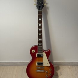 Elektrická kytara s trupem v červeně-žlutém sunburst provedení a bílým pickguardem. Na hlavě hmatníku je bílý nápis Epiphone a pod ním kurzivou Les P…