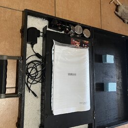 Otevřený černý kufřík s bílou vnitřní výplní, ve kterém je umístěno černé zařízení a dokumentace v plastovém obalu s nápisem YAMAHA. K zařízení je př…