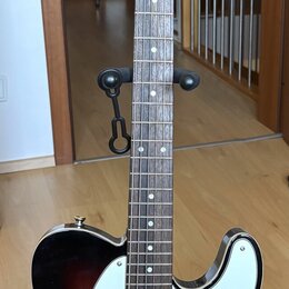 Elektrická kytara s tmelelovým korpusem v barevném přechodu od černé k hnědé a bílým pickguardem. Na hlavě hmatníku je viditelné logo Squier. Hmatník…