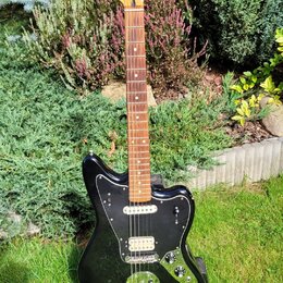Černá elektrická kytara s černým pickguardem a světle hnědým hmatníkem. Na hlavě je viditelné logo Fender a nápis Japan. Nástroj má dva snímače, tři…