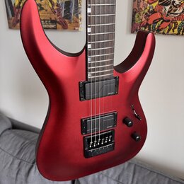 Elektrická kytara s červeným matným povrchem a hnědým hmatníkem. Nástroj má dva černé humbuckery a černý most s integrovaným pákovým systémem. Na těl…
