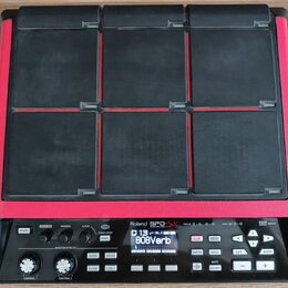 Černý sampler s devíti gumovými pady a červeným rámem. Na spodním ovládacím panelu je bílý nápis Roland SP-90 SX. Zařízení obsahuje displej s textem…