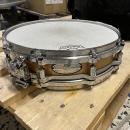Na fotce je malý snare drum s dřevěným korpusem v přirozené hnědé barvě a chromovaným kováním. Na korpusu je viditelný oválný štítek s nápisy „Pearl“…