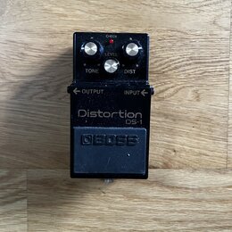 Černý efekt pedál s nápisy Distortion a DS-1. Na horní části jsou tři stříbrné ovládací knoflíky s popisky LEVEL, TONE a DIST, červená kontrolka CHEC…