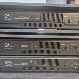Tři černé rackové výkonové zesilovače značky QSC AUDIO. Horní jednotka nese nápis RMX 1850HD PROFESSIONAL POWER AMPLIFIER, prostřední RMX 1450 PROFES…